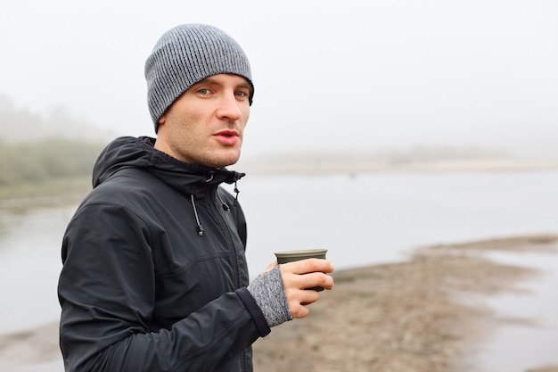 Hombre guapo con chaqueta negra y gorra gris sosteniendo una taza de metal con té caliente contra el río brumoso