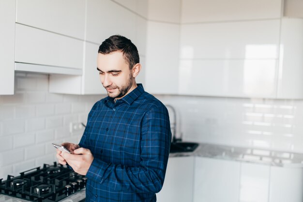 Hombre guapo en la cocina está sonriendo por la mañana y mirando el teléfono inteligente