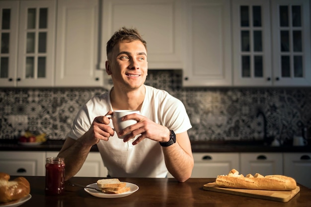 Un hombre guapo en la cocina sonríe y bebe café por la mañana