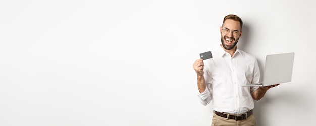 Hombre guapo de compras en línea que muestra la tarjeta de crédito y usa una computadora portátil para ordenar en internet de pie