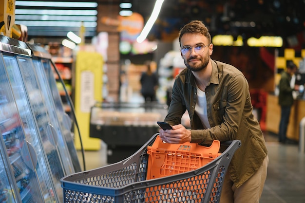 Hombre guapo de compras en el supermercado sonriendo usando el teléfono