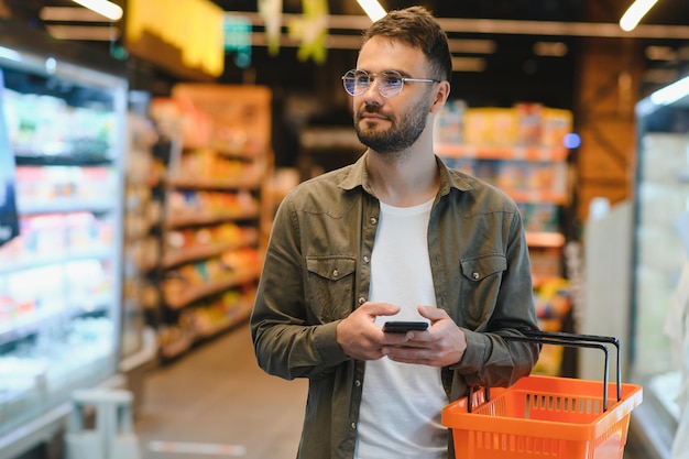 Hombre guapo de compras en el supermercado sonriendo usando el teléfono