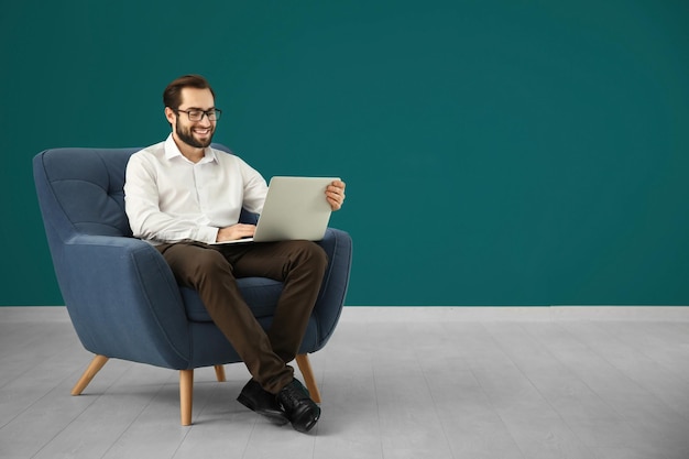 Hombre guapo con computadora portátil sentado en un cómodo sillón contra la pared de color