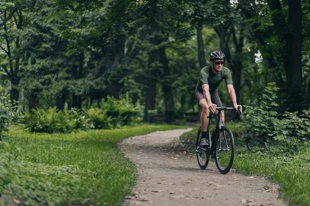 Hombre guapo con cuerpo musculoso montando bicicleta negra en la carretera.  chico caucásico sano y activo en casco y gafas de entrenamiento al aire  libre. concepto de ciclismo de pista. | Foto