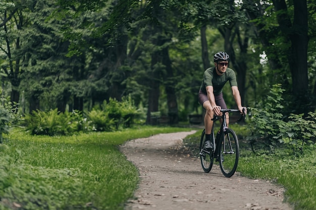 Hombre guapo con cuerpo musculoso montando bicicleta negra en la carretera. Chico caucásico sano y activo en casco y gafas de entrenamiento al aire libre. Concepto de ciclismo de pista.