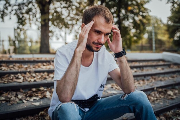 Un hombre guapo en depresión Hipster no afeitado se sienta en los escalones en el parque de otoño sosteniendo su cabeza Concepto el dolor de cabeza y el dolor