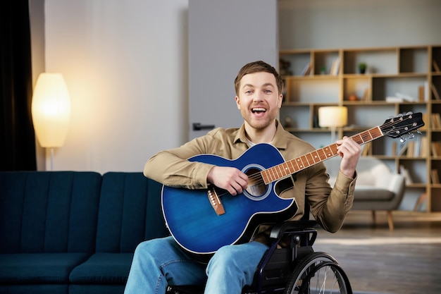 Hombre guapo con discapacidad sentado en silla de ruedas tocando la guitarra en casa