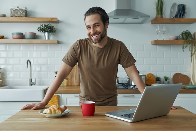 Hombre guapo disfrutando del desayuno y mirando a la cámara mientras pasa tiempo en la cocina