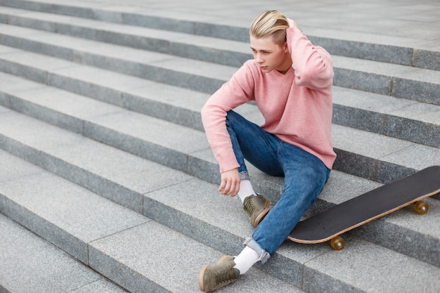 Hombre guapo elegante en ropa de moda con una patineta se sienta en los escalones