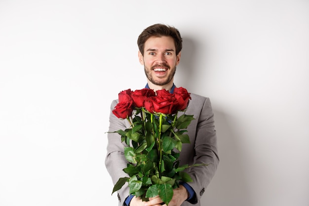 Hombre guapo enamorado deseando feliz día de San Valentín, dando ramo de flores en una cita romántica, sonriendo a la cámara, vestido con traje sobre fondo blanco.