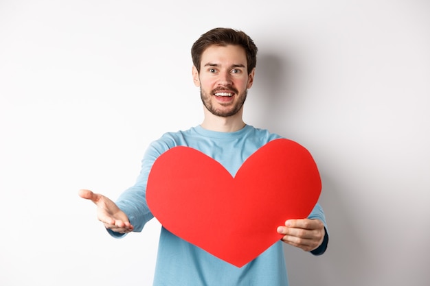 Hombre guapo enamorado haciendo una confesión, apuntando con la mano a la cámara, sosteniendo un gran corazón rojo en el día de San Valentín, cantando una serenata romántica, de pie sobre un fondo blanco.