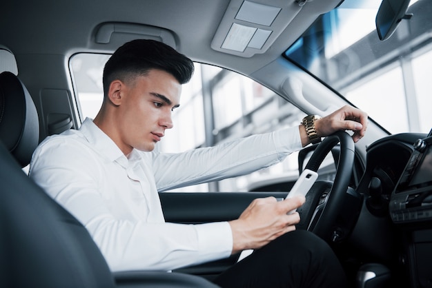 El hombre guapo es un comprador sentado en un automóvil con un teléfono en un automóvil nuevo en un centro de distribución.