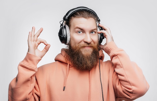 Un hombre guapo escandinavo muestra un gesto correcto, sonríe y escucha música en auriculares profesionales aislados en un fondo gris. Chico feliz con barba y peinado de jengibre. Tecnologías digitales modernas.