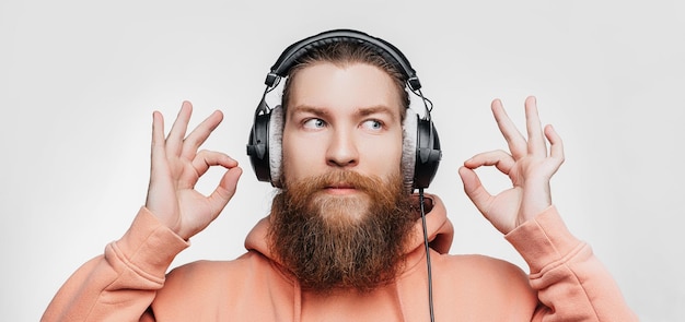 Hombre guapo escandinavo muestra gesto ok mantener la calma y escucha música en auriculares profesionales aislados sobre fondo gris. Chico feliz con barba y peinado de jengibre. Tecnologías digitales modernas