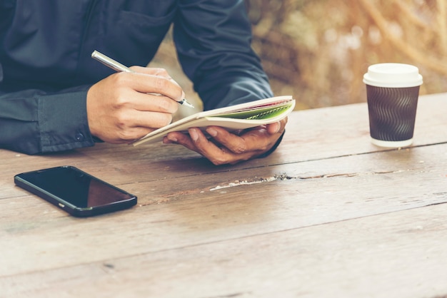 Hombre guapo escribir diario de nota sobre la mesa de madera con teléfono móvil y taza de café.
