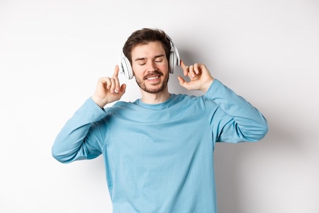 Hombre guapo escuchando música en auriculares inalámbricos y sonriendo, disfrutando de un buen sonido, fondo blanco.