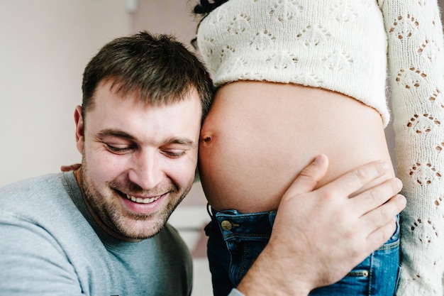 Hombre guapo está escuchando la barriga de su hermosa esposa embarazada y sonriendo Mujer y hombre amoroso abrazando la barriga en casa Concepto de paternidad de pareja amorosa Baby Shower