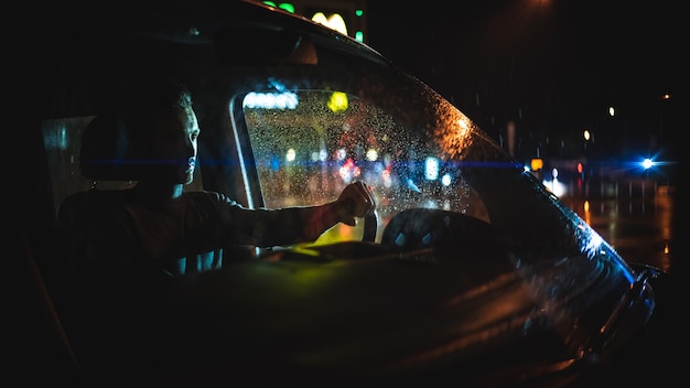 El hombre guapo está sentado en el coche en la calle lluviosa de la noche.