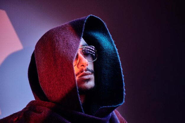 Hombre guapo con estilo en capucha y gafas de sol posando en el estudio bajo luces azules y rojas.