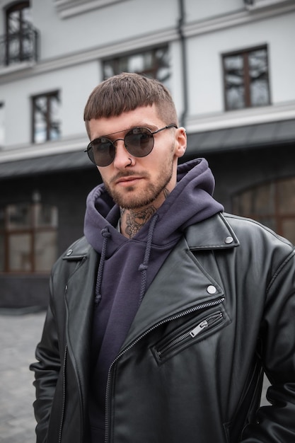 Hombre guapo con estilo hipster con gafas de sol de moda en ropa de moda con chaqueta de cuero y sudadera con capucha caminando en la ciudad