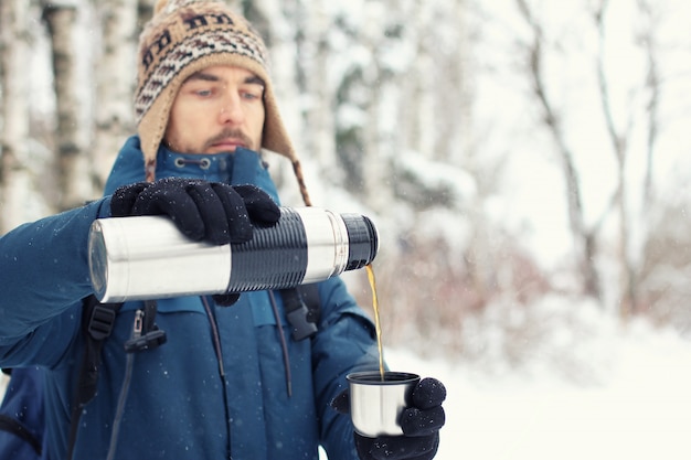 Hombre guapo excursionista en ropa de abrigo vierte té en la taza del termo en bosque de invierno.