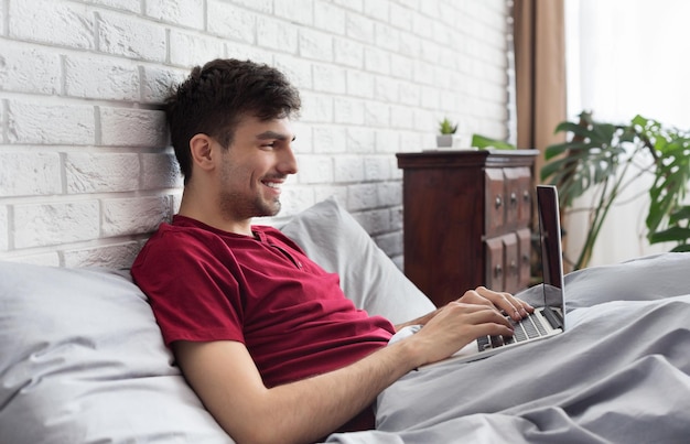 Foto hombre guapo feliz usando laptop en la cama por la mañana