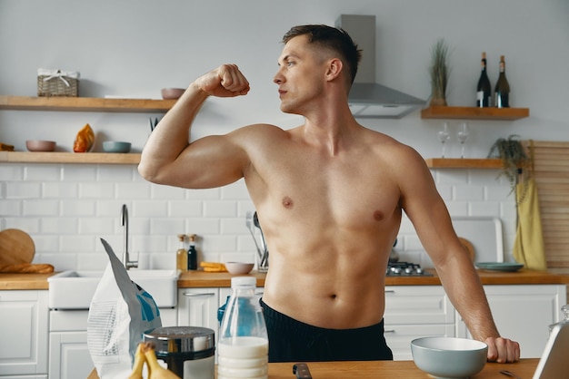 Foto hombre guapo y en forma mostrando su bíceps perfecto mientras prepara comida saludable en la cocina