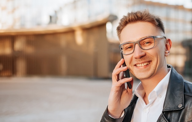 Hombre guapo con gafas con un teléfono inteligente en la calle de una gran ciudad. Empresario hablando por teléfono sobre fondo urbano
