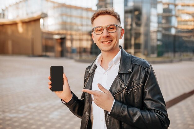 Hombre guapo con gafas con un teléfono inteligente en la calle de una gran ciudad Empresario señala con el dedo la pantalla del teléfono en el fondo urbano