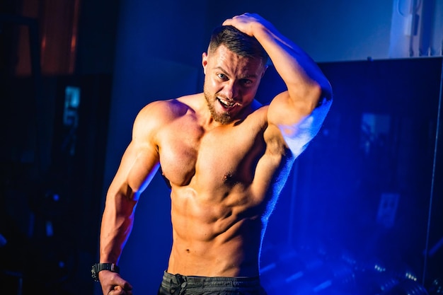 Hombre guapo con grandes músculos posando en la cámara en el gimnasio Fondo negro y azul Retrato de un culturista sonriente Primer plano