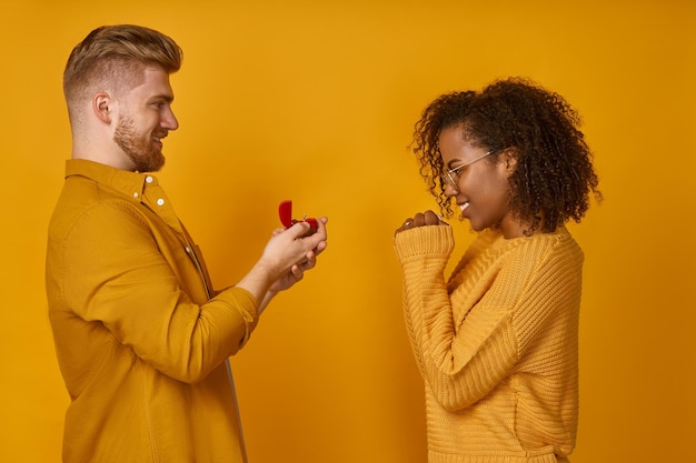 Hombre guapo haciendo una propuesta a la feliz mujer de piel oscura con caja de anillo