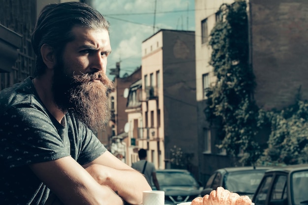 Hombre guapo hipster con barba larga y bigote con croissant y taza de café en la cafetería al aire libre en