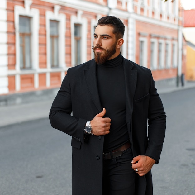 Hombre guapo hipster de moda con barba y bigote en un abrigo negro en la calle