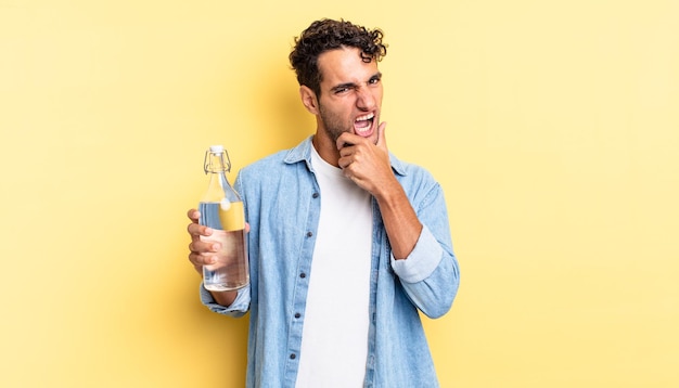 Hombre guapo hispano con la boca y los ojos bien abiertos y la mano en la barbilla. concepto de botella de agua