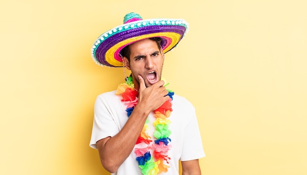 Hombre guapo hispano con la boca y los ojos bien abiertos y la mano en la barbilla. concepto de fiesta mexicana