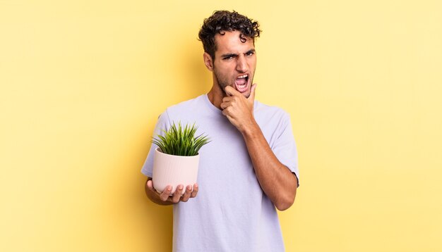 Hombre guapo hispano con la boca y los ojos bien abiertos y la mano en la barbilla. concepto de planta decorativa