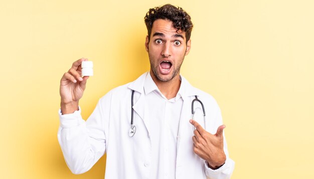 Hombre guapo hispano mirando consternado y sorprendido con la boca abierta, apuntando a sí mismo. concepto de píldoras de botella de médico