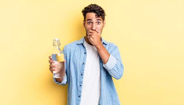 Hombre guapo hispano que cubre la boca con las manos con una sorpresa. concepto de botella de agua