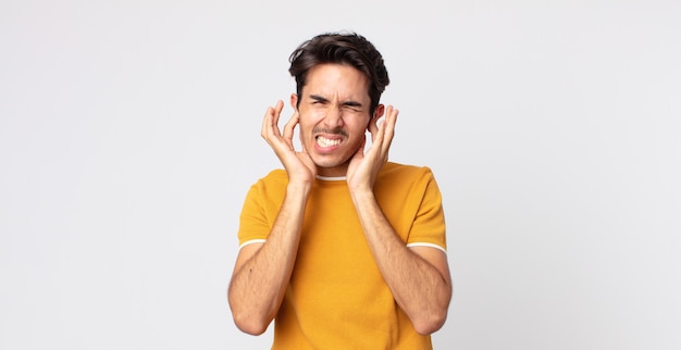 Hombre guapo hispano que parece enojado, estresado y molesto, cubriendo ambos oídos con un ruido ensordecedor, sonido o música alta