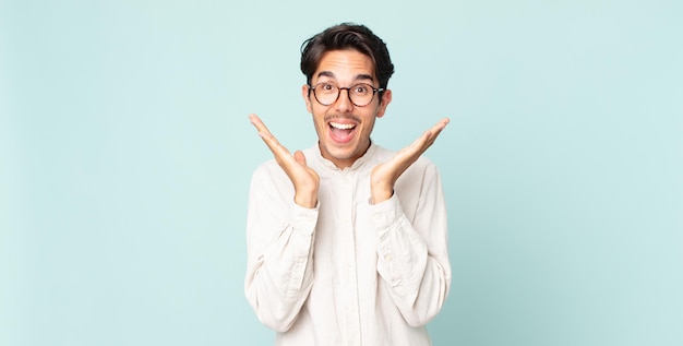 Hombre guapo hispano que parece feliz y emocionado, sorprendido con una sorpresa inesperada con ambas manos abiertas junto a la cara