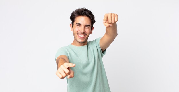 Hombre guapo hispano que se siente feliz y confiado, apuntando a la cámara con ambas manos y riendo, eligiéndote