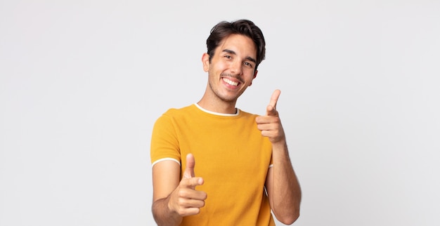 Hombre guapo hispano sonriendo con una actitud positiva, exitosa y feliz apuntando a la cámara, haciendo un signo de pistola con las manos