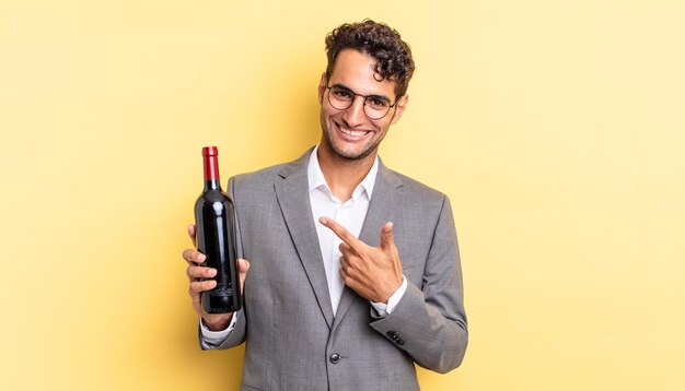 Hombre guapo hispano sonriendo alegremente sintiéndose feliz y señalando el concepto de botella de vino lateral