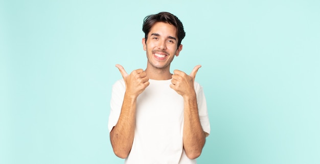 Hombre guapo hispano sonriendo con alegría y luciendo feliz, sintiéndose despreocupado y positivo con ambos pulgares hacia arriba