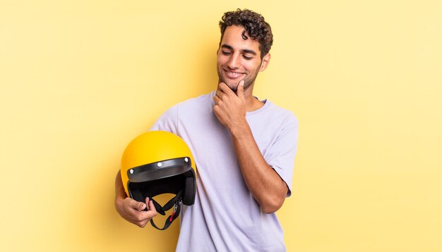 Hombre guapo hispano sonriendo con una expresión feliz y segura con la mano en la barbilla. concepto de casco de moto