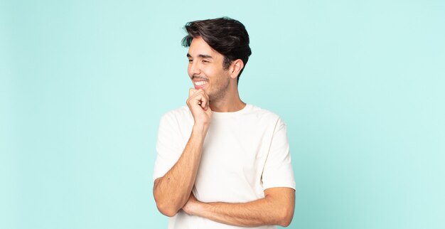 Hombre guapo hispano sonriendo con una expresión feliz y segura con la mano en la barbilla, preguntándose y mirando hacia un lado