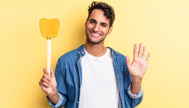 Hombre guapo hispano sonriendo felizmente, saludando con la mano, dándote la bienvenida y saludándote. mata moscas concepto