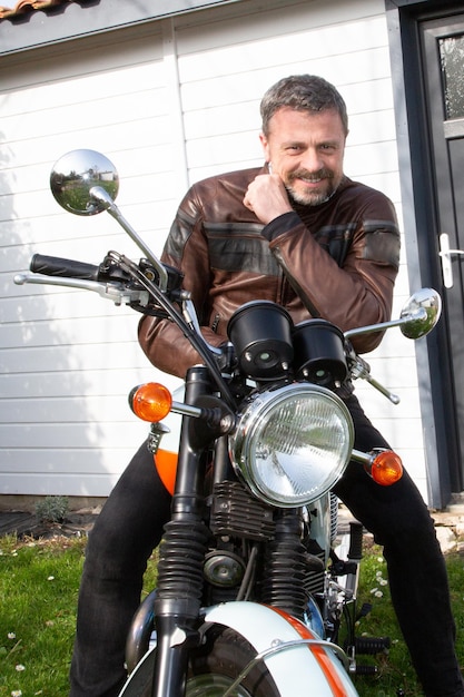 Hombre guapo jinete con barba en chaqueta de motociclista de cuero marrón a un lado moto retro vintage neo