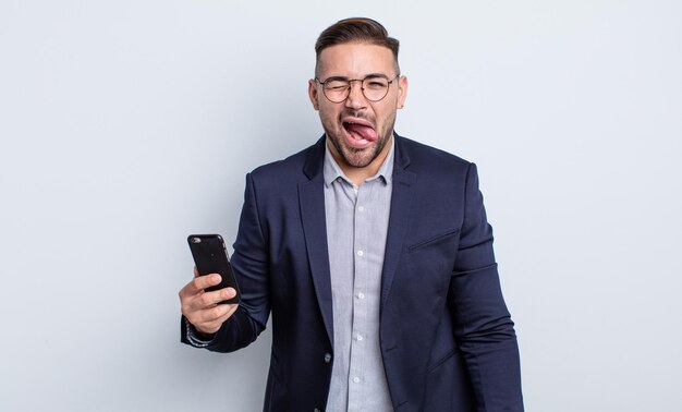 Hombre guapo joven con actitud alegre y rebelde, bromeando y sacando la lengua concepto de negocio y teléfono inteligente