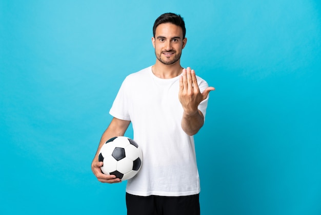 Hombre guapo joven aislado en la pared azul con balón de fútbol y haciendo el gesto que viene